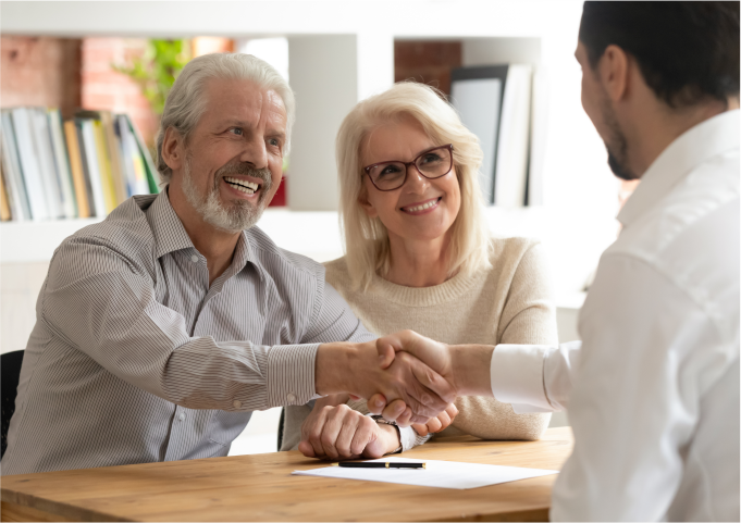 A smiling elderly couple shaking hands with a professional, symbolizing trust and successful agreement in a professional office.